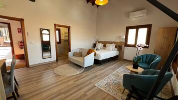Una habitación amplia con una cama grande, una zona de estar con un sofá y sillones, una mesita con un florero y un techo de madera.
