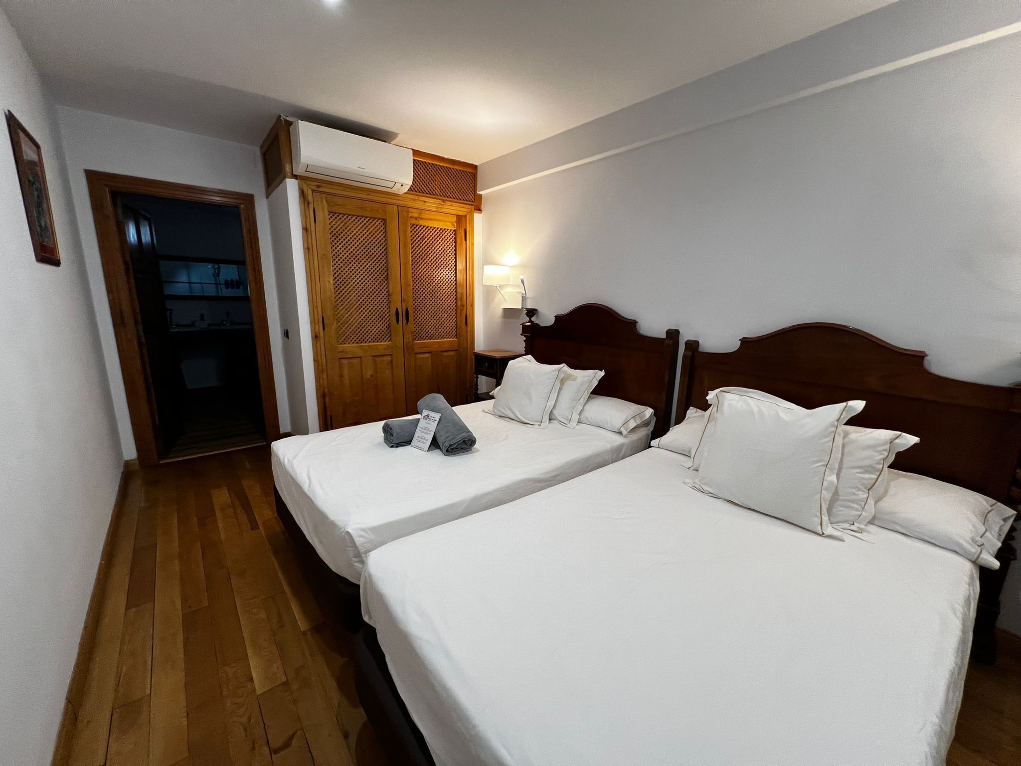 A hotel room with two beds, wooden wardrobe, and a ceiling air conditioner.