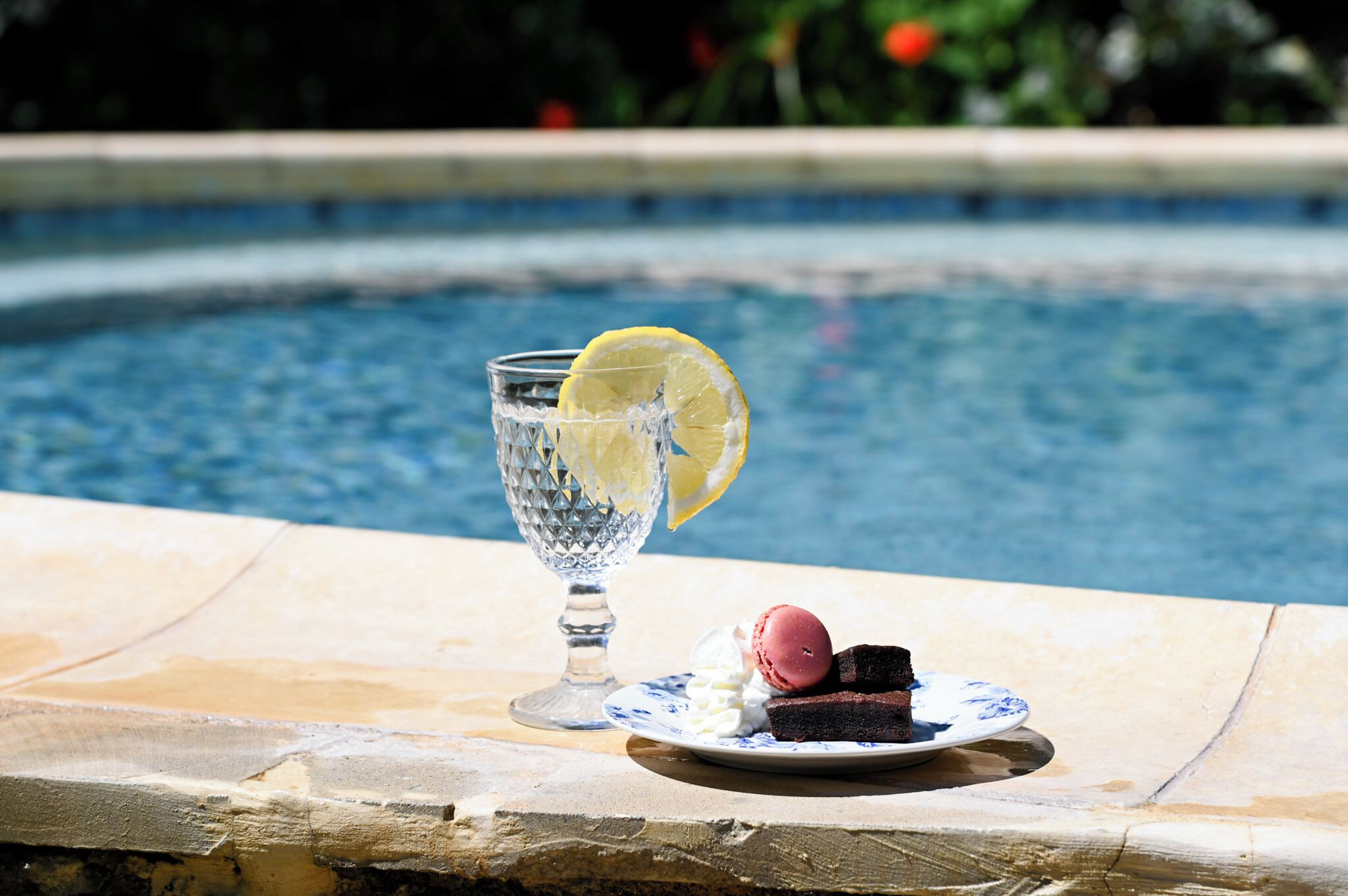 A glass of water with a lemon slice, a plate with a dessert, and a pool in the background.