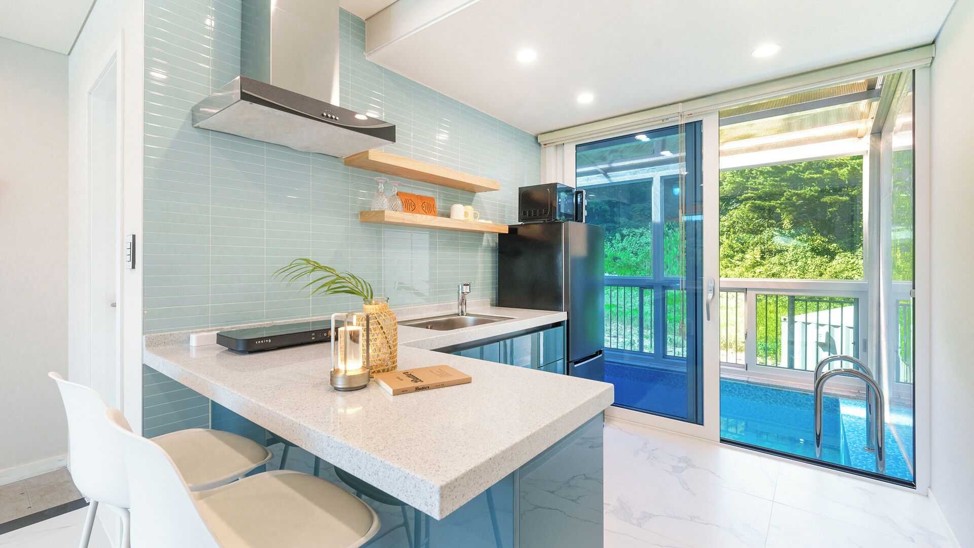 A modern kitchen with a built-in stove, a microwave, and a refrigerator. There is a window with a view of a pool and greenery.