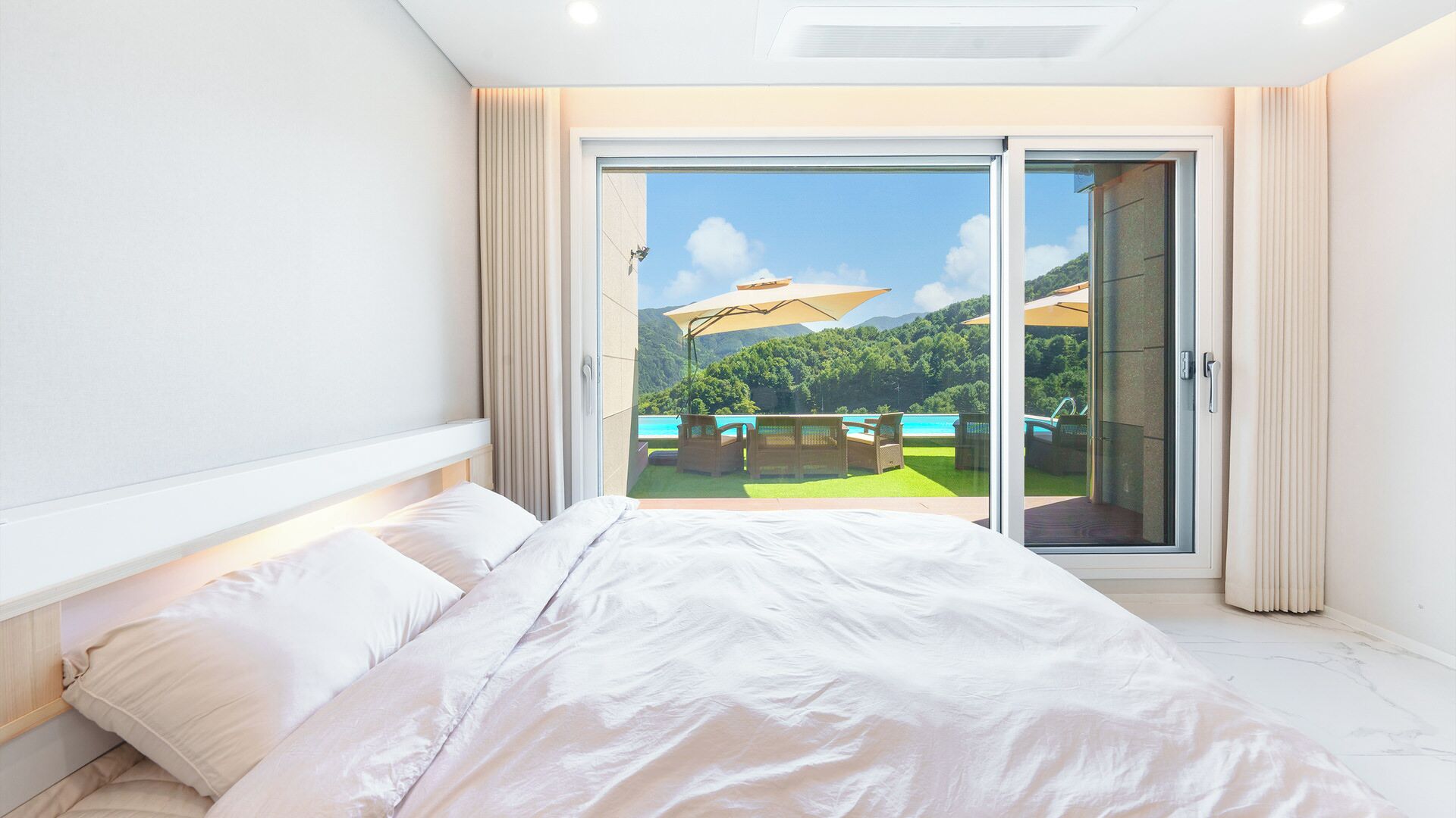 A bedroom with a large bed, a sliding glass door leading to a balcony with outdoor furniture and a view of a mountainous landscape.