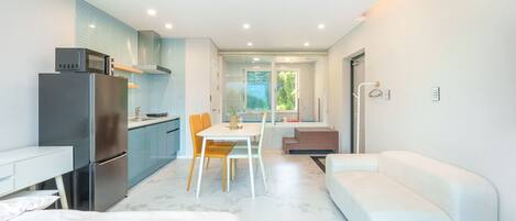 A modern kitchen with a refrigerator, stove, and sink. There is a dining area with a table and chairs. A white sofa and a small couch are in the living area.