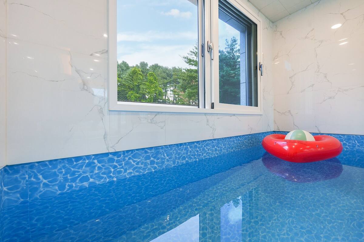 Une piscine dont le bord est orné de mosaïques bleues, une fenêtre donnant sur les arbres et un flotteur rouge gonflable.