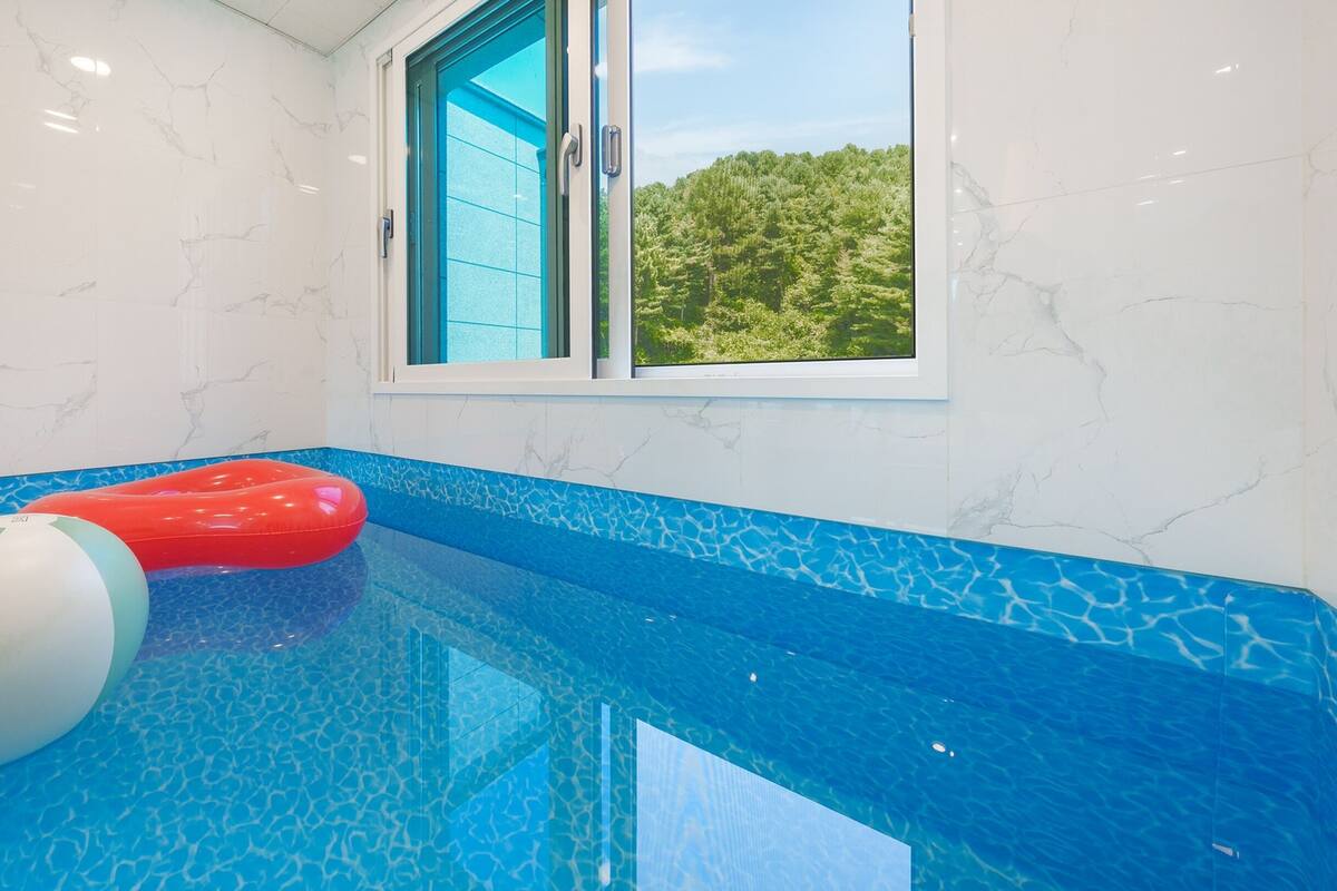 Une piscine dont le sol est recouvert de mosaïques bleues, un ballon gonflable rouge et blanc, et une fenêtre donnant sur des arbres verdoyants.