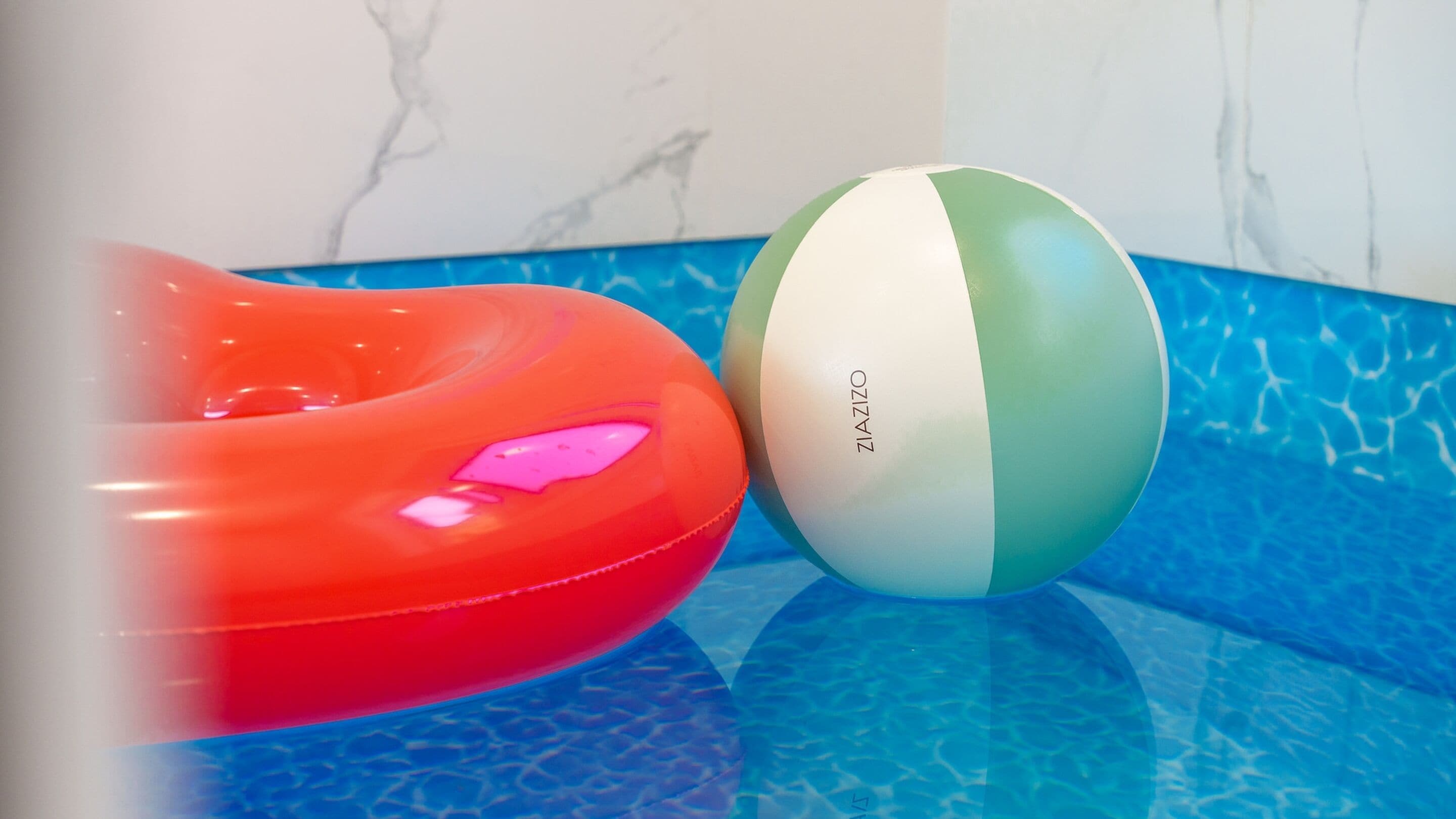 A pool with a blue and white tiled floor, a red inflatable ring, and a white and green beach ball.