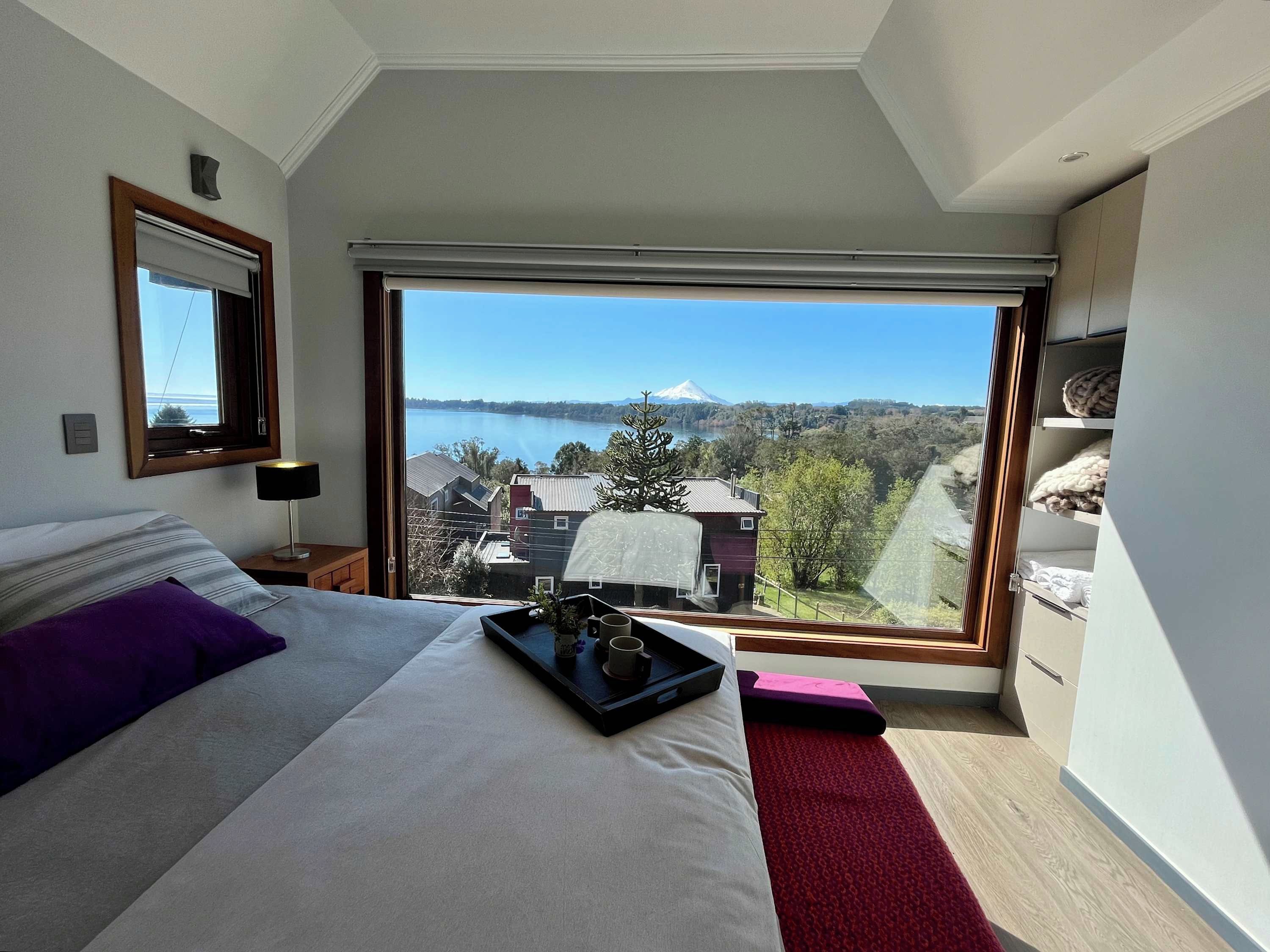 A bedroom with a large window, a bed, a bedside table, and a view of a lake and mountains.