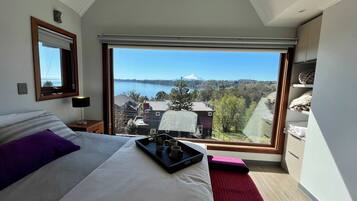 A bedroom with a large window, a bed, a bedside table, and a view of a lake and mountains.