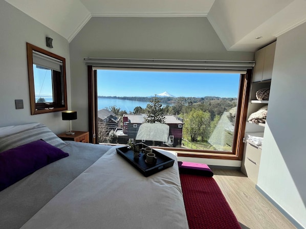 Un dormitorio con un ventanal grande, una cama, una mesita de noche y vistas a un lago y montañas.
