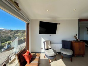 Ein Wohnzimmer mit großem Fenster, einem Flachbildfernseher, einer Couch, zwei Sesseln und einer Holzkommode.