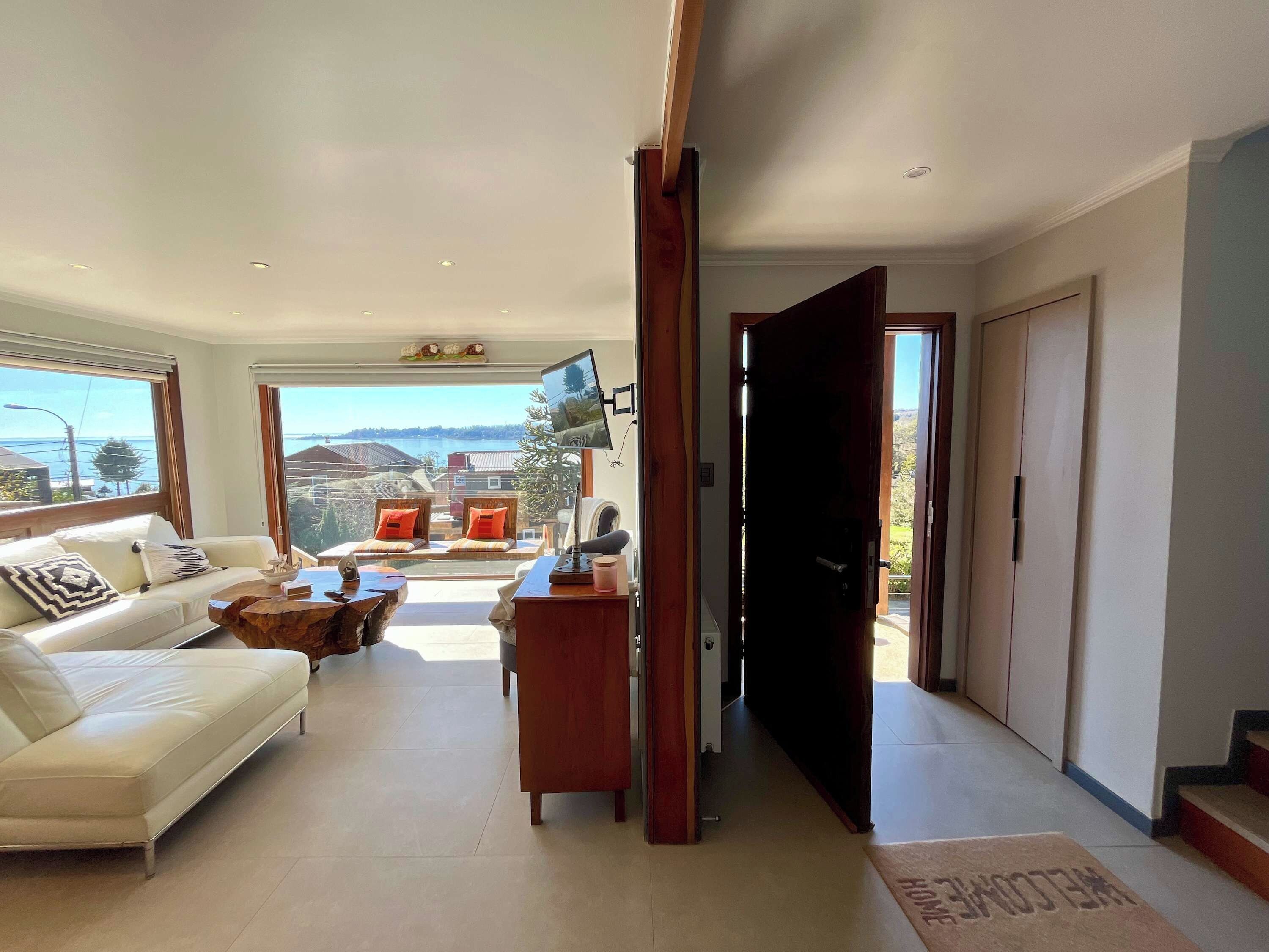 A spacious living area with a sofa, coffee table, and a TV mounted on the wall. There is a view of the outdoors through large windows.