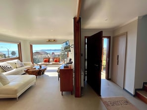 Ein großzügiger Wohnbereich mit einer Couch, einem Couchtisch und einem an der Wand befestigten Fernseher. Große Fenster bieten Blick ins Freie.