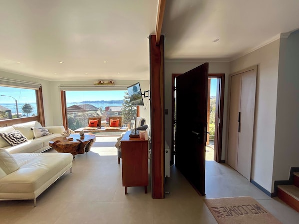 A spacious living area with a sofa, coffee table, and a TV mounted on the wall. There is a view of the outdoors through large windows.