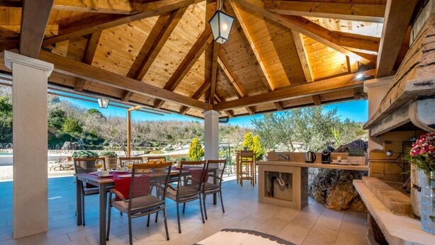 A covered outdoor area with a wooden ceiling, a table with chairs, and a stone fireplace. - Villa Pizzelli (Dubrovnik)