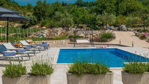 A rectangular swimming pool with lounge chairs and umbrellas, surrounded by greenery and a rocky landscape. - Villa Pizzelli (Dubrovnik)