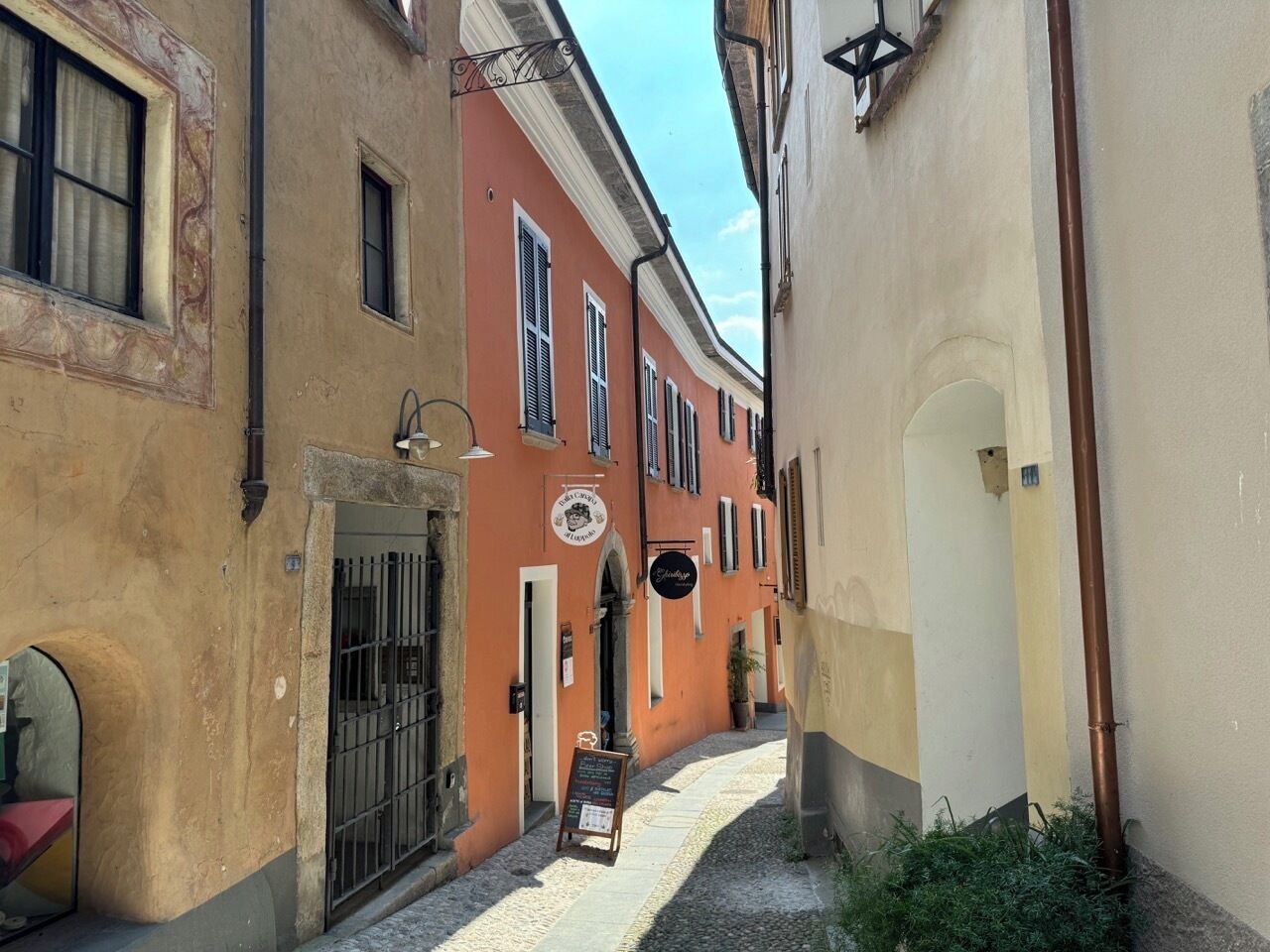 A narrow street with buildings on either side, a signboard, and a small garden area.