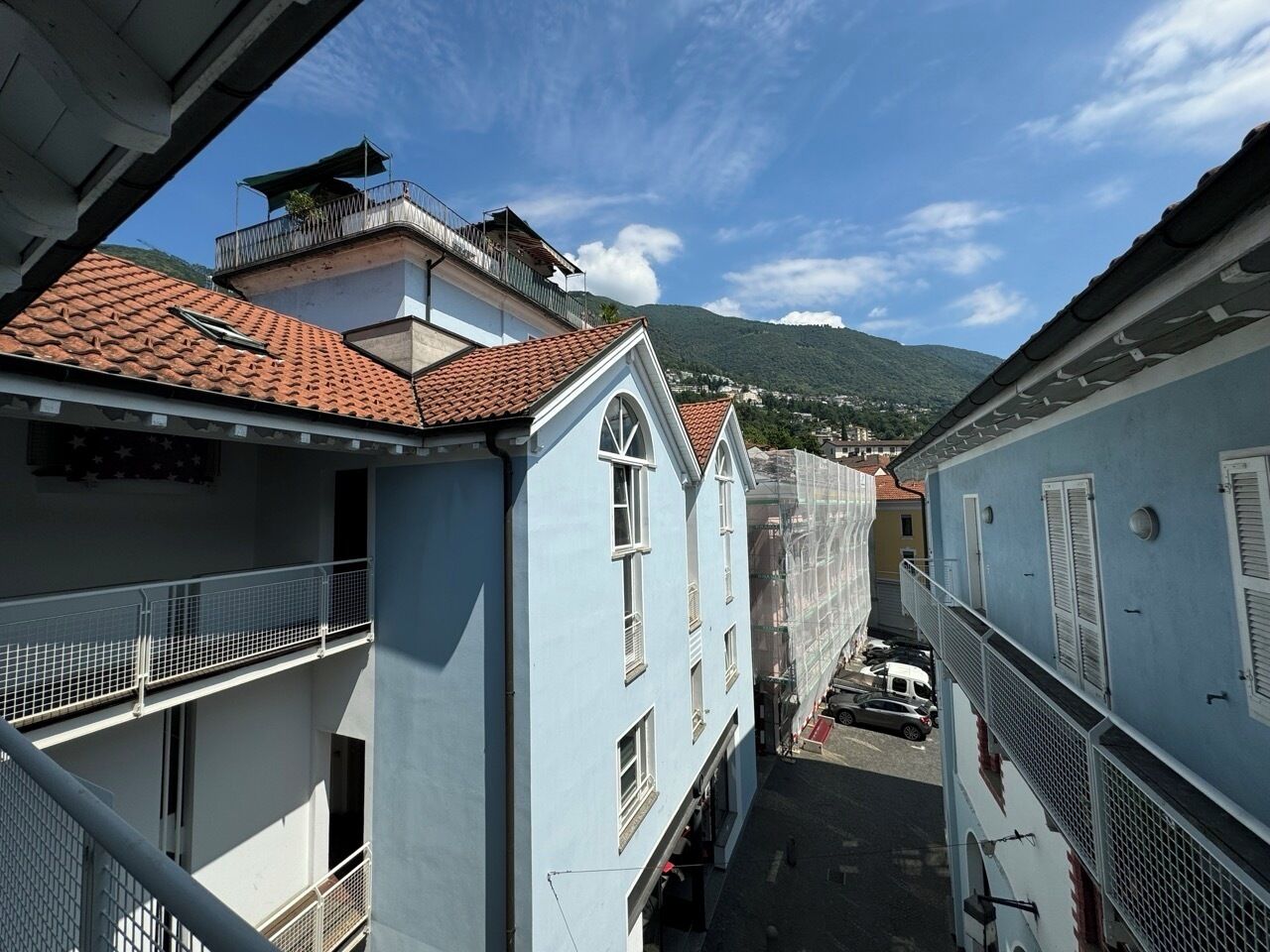 A narrow street with buildings on either side, featuring balconies and a mountain in the background.