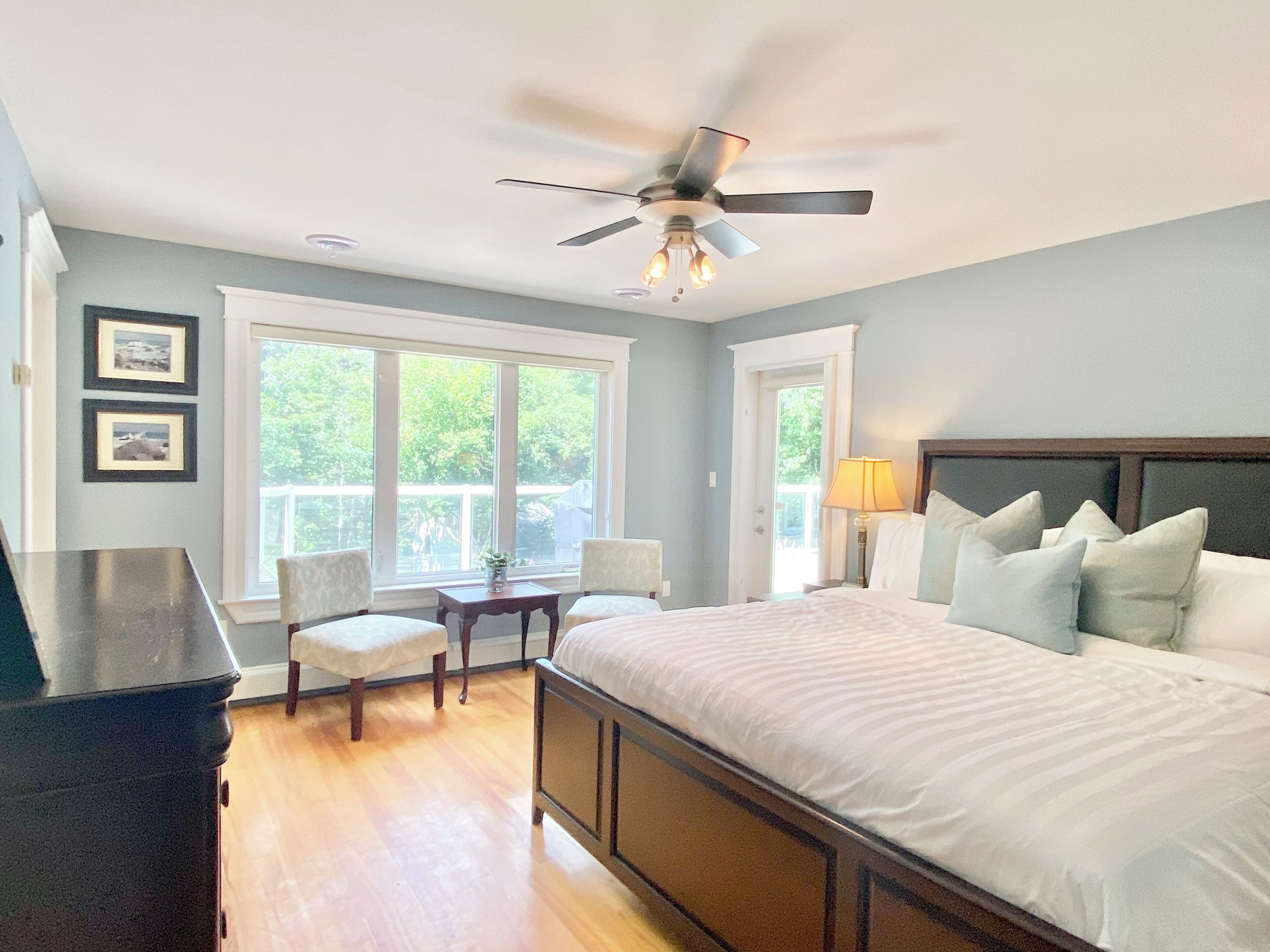 A bedroom with a large bed, a ceiling fan, a desk, and two chairs. There are two windows with white curtains and a view of greenery outside.