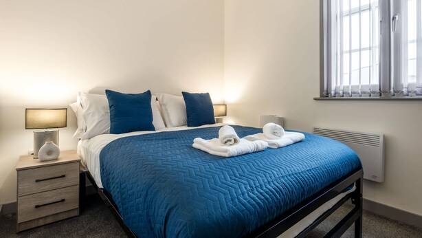 A neatly made bed with a blue quilt, two pillows, and two towels folded on top. There is a bedside lamp on the left and a window with blinds on the right.
