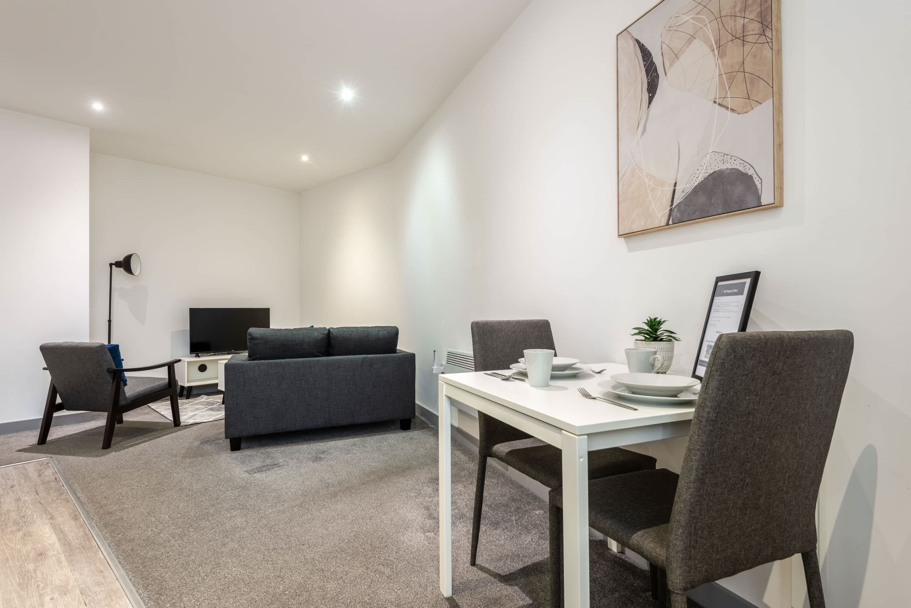 Une pièce moderne et bien éclairée, avec une table blanche dressée pour le repas, un canapé gris et un téléviseur en arrière-plan.