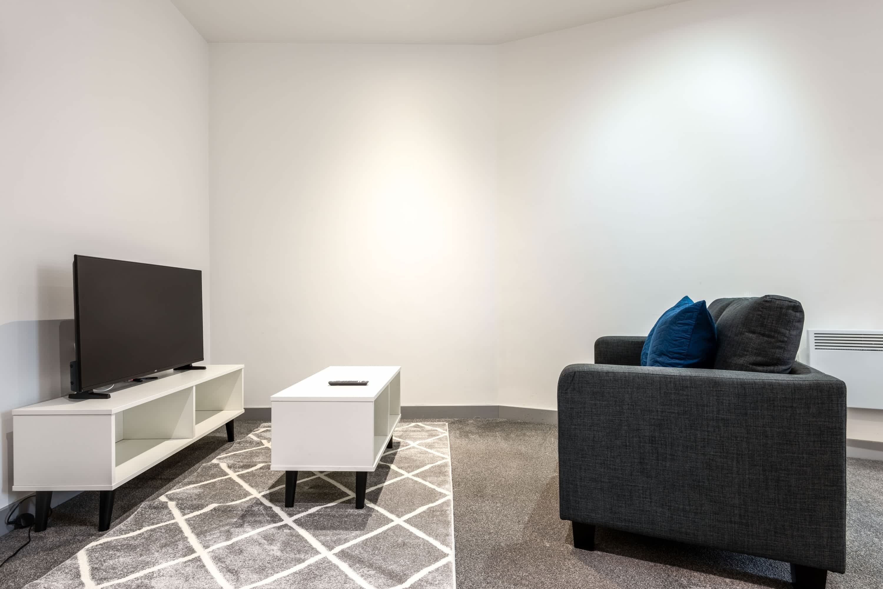 A modern living room with a flat-screen TV on a white stand, a small white side table, and a grey sofa with blue cushions.