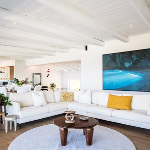 Een ruime woonkamer met een witte modulaire bank, een ronde salontafel en een groot schilderij van een blauw landschap.