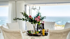 A dining table set with chairs, a vase of flowers, and a view of the sea.