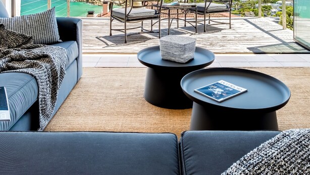 A balcony with a sofa, two round tables, and a view of the ocean and rocky coastline.