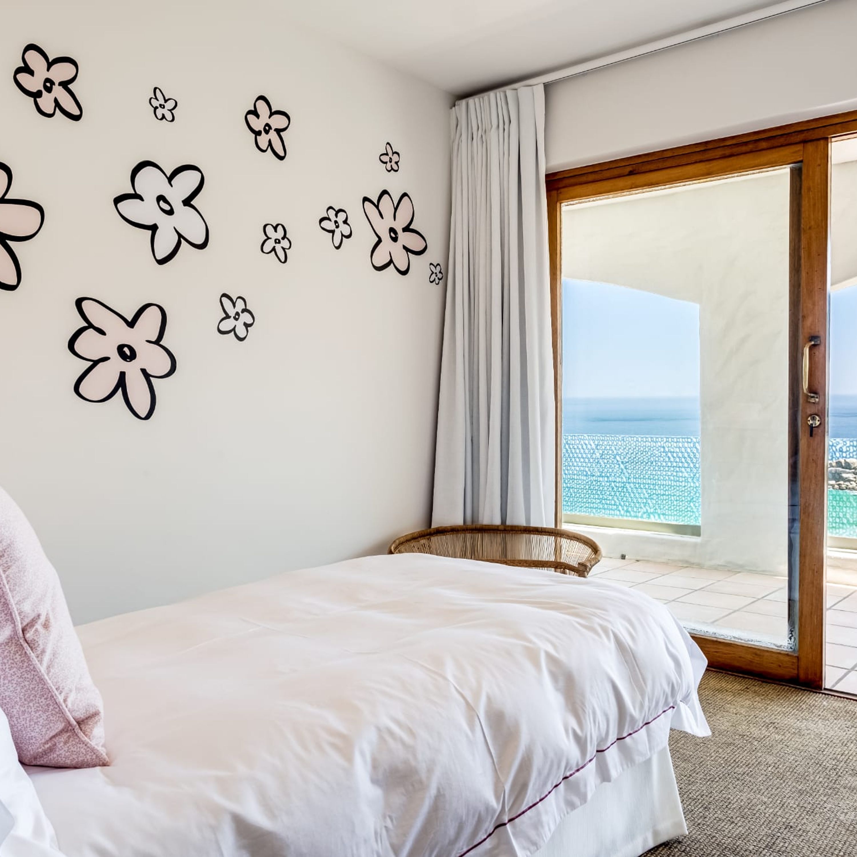Un dormitorio con una cama, una silla de mimbre, vistas al mar y diseños florales en la pared.
