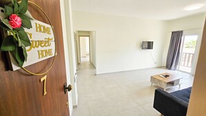Un vaste espace de vie agrémenté d’une porte en bois, d’une enseigne portant l’inscription « Chez soi, quel bonheur ! », d’un téléviseur à écran plat fixé au mur et d’une porte-fenêtre coulissante donnant sur un espace extérieur.