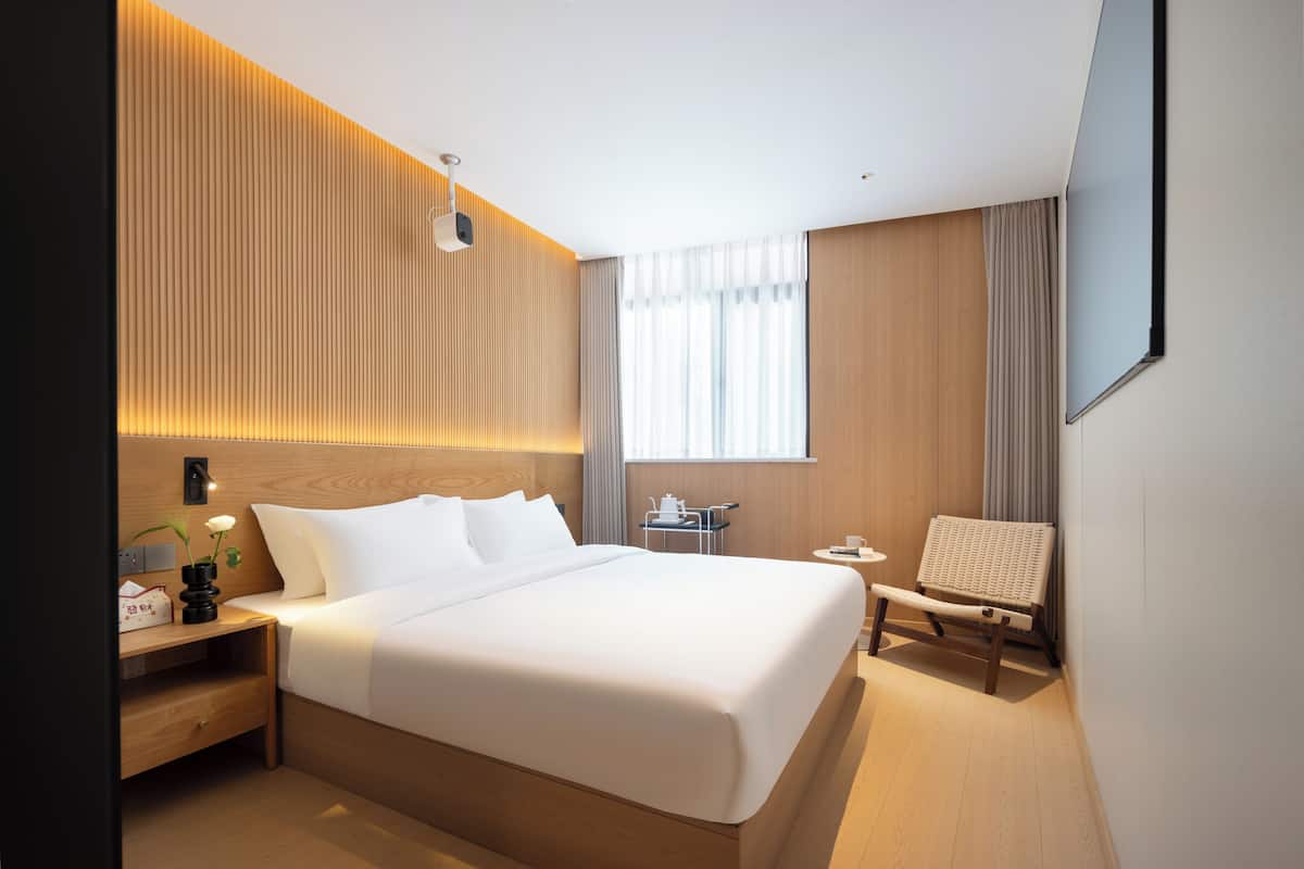 Ein modernes Hotelzimmer mit einem großen Bett, einem Nachttisch, einem kleinen Schreibtisch, einem Sessel und einem Fenster mit Vorhängen.