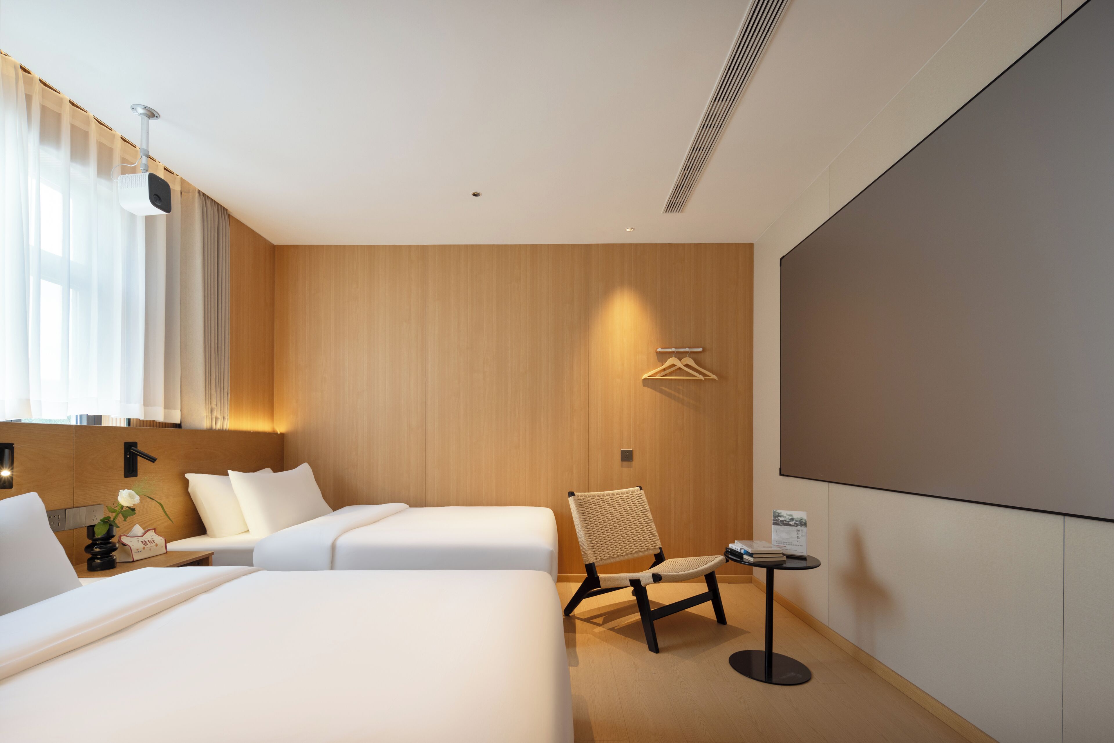 Een moderne hotelkamer met twee bedden, een zithoek met een stoel en een tafel, een scherm voor een beamer, en een houten wand.