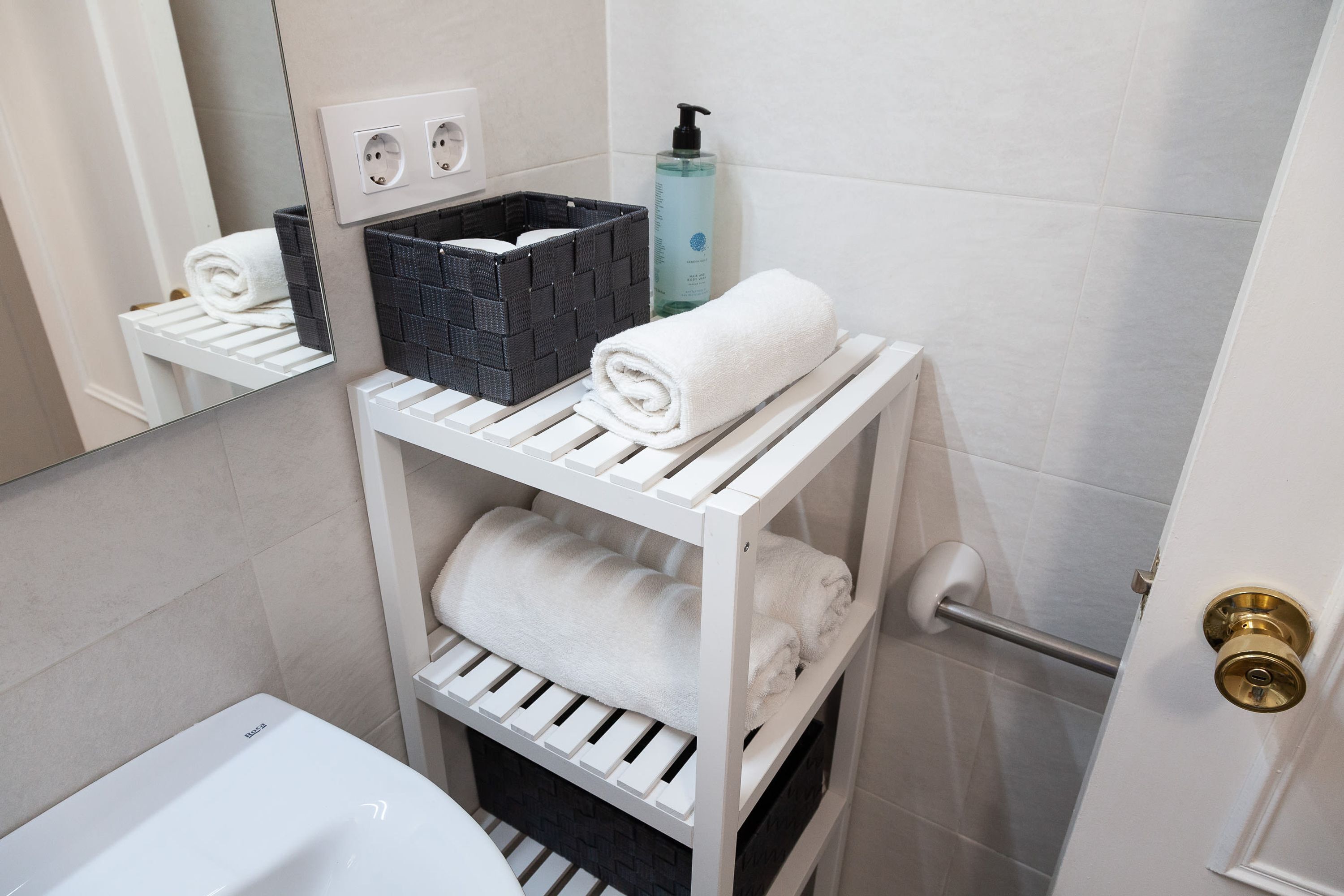 A bathroom with a white sink, a towel rack with folded towels, a black basket, and a bottle of liquid soap.