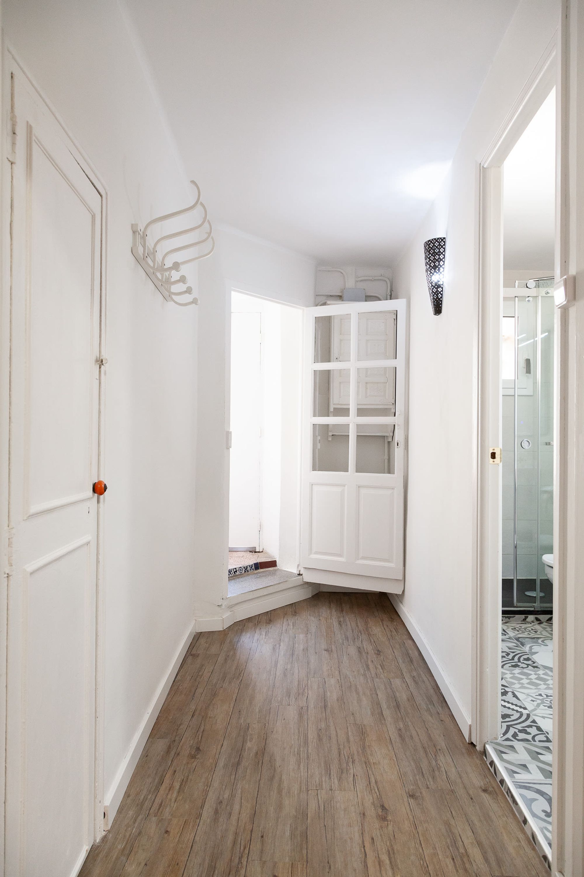 A narrow hallway with wooden flooring, white walls, and a white door with an orange handle.