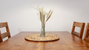 Ein Holztisch mit einer gewebten Tischmatte, eine transparente Vase mit getrockneten Weidengräsern und zwei Holzstühle.