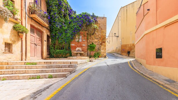 Eine enge Gasse mit einem Steinfbrunnen, Stufen, die zu einem Gebäude mit einer Holztür führen, und eine gelbe Mauer auf der rechten Seite.