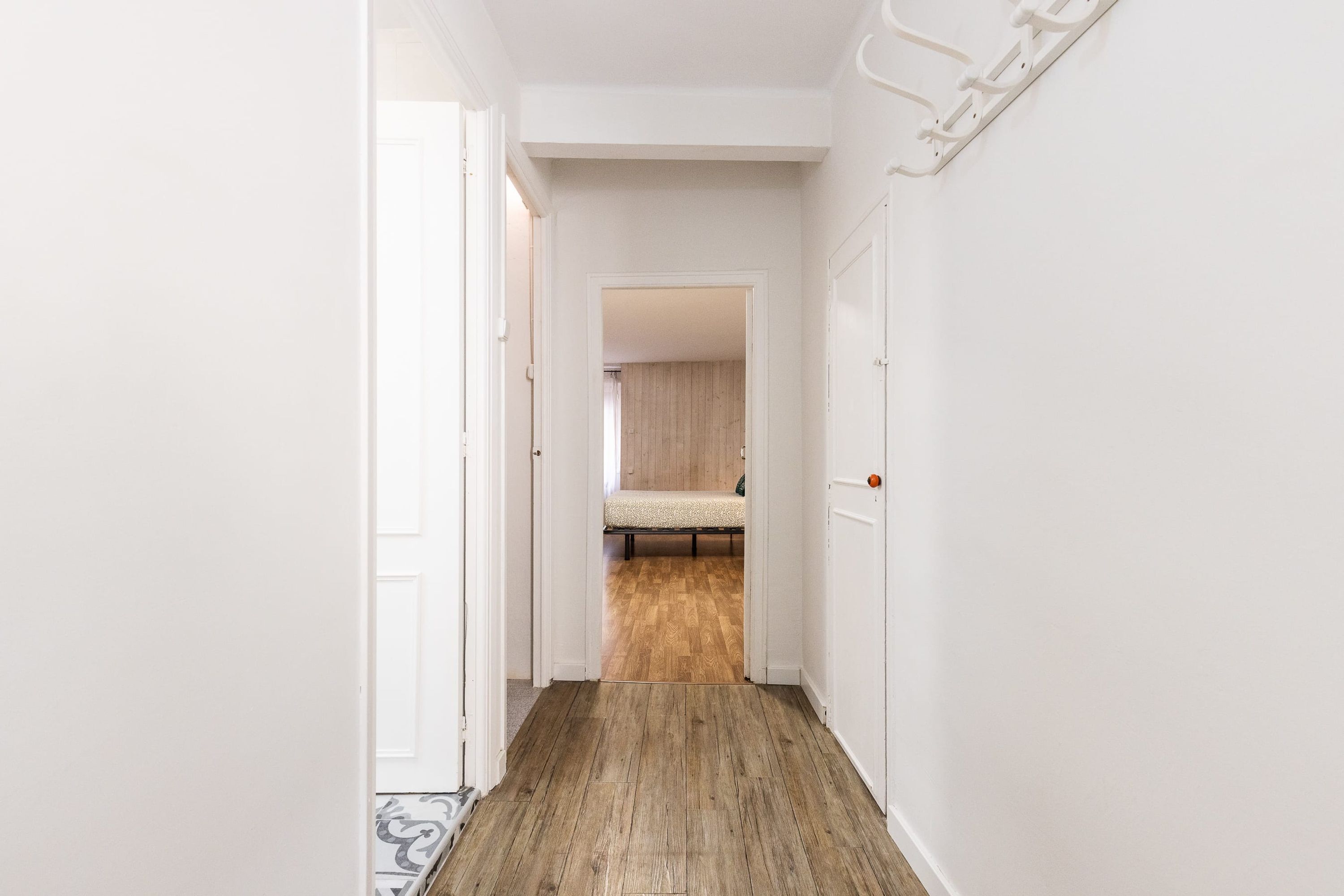 A narrow hallway with wooden flooring leading to a bedroom with a bed and a wooden floor.