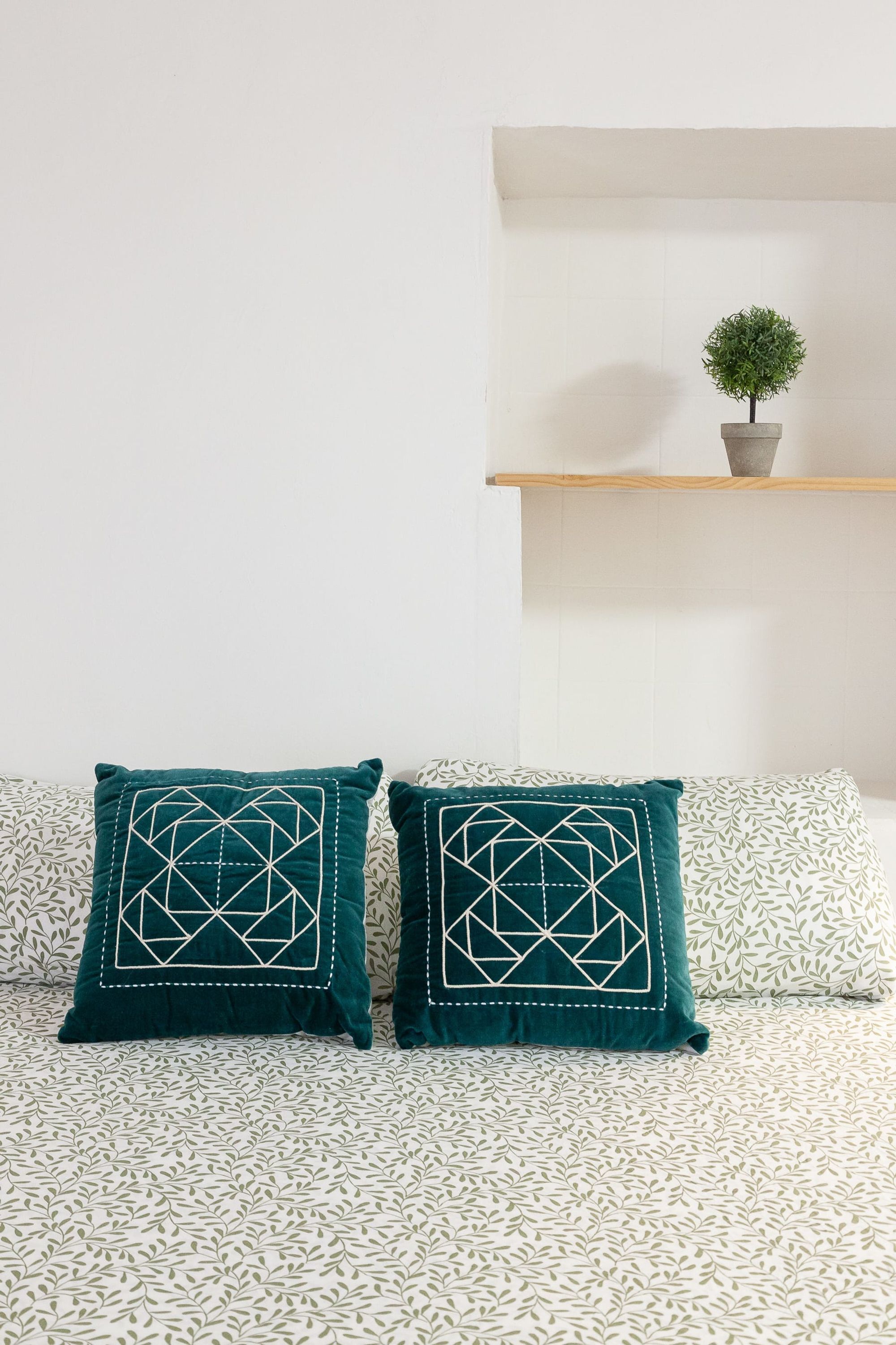 Una cama con ropa de cama estampada, dos almohadas turquesas con diseños geométricos y una pequeña planta en maceta sobre un estante.