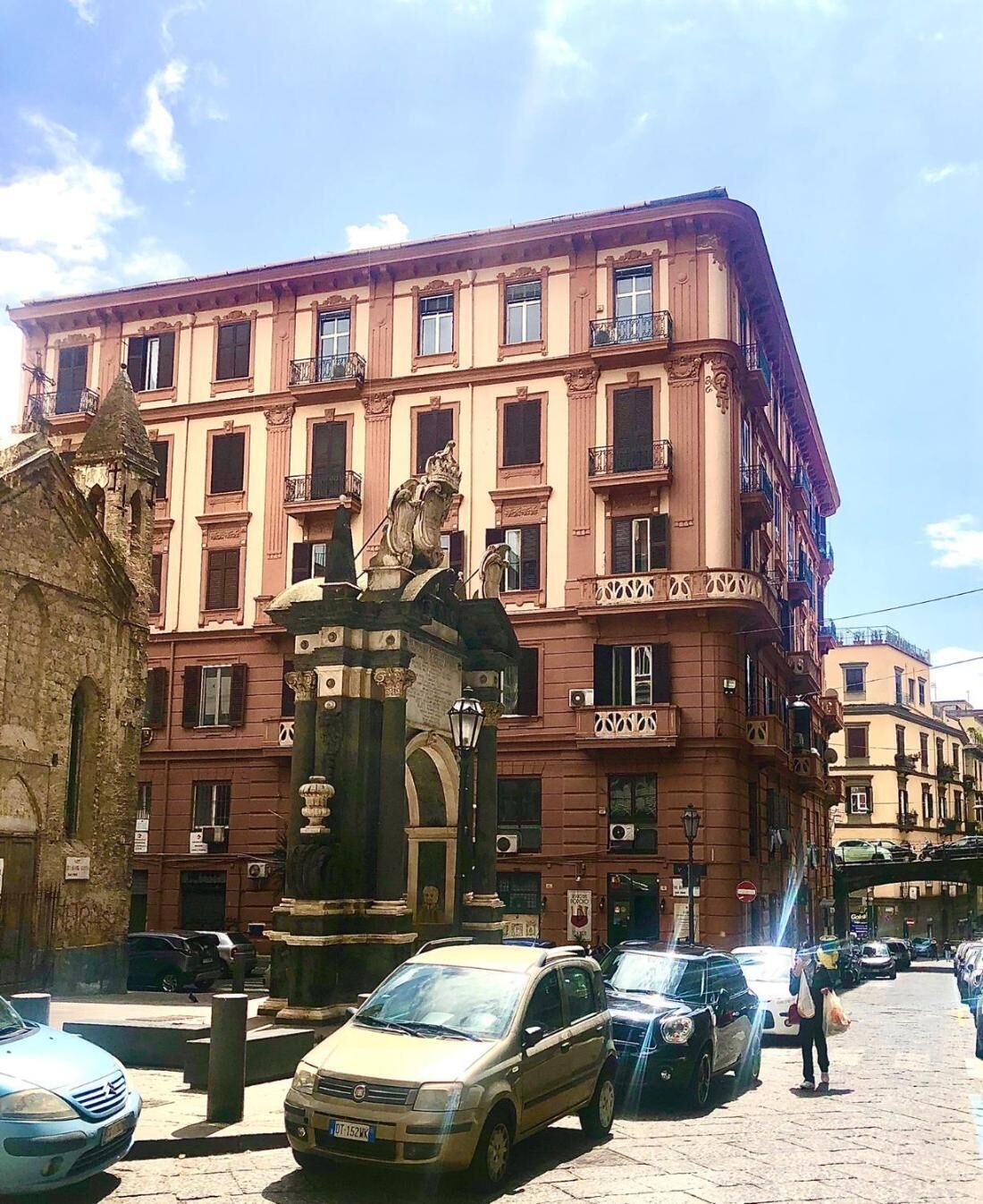 A multi-story building with a statue in front, parked cars, and a cobblestone street.