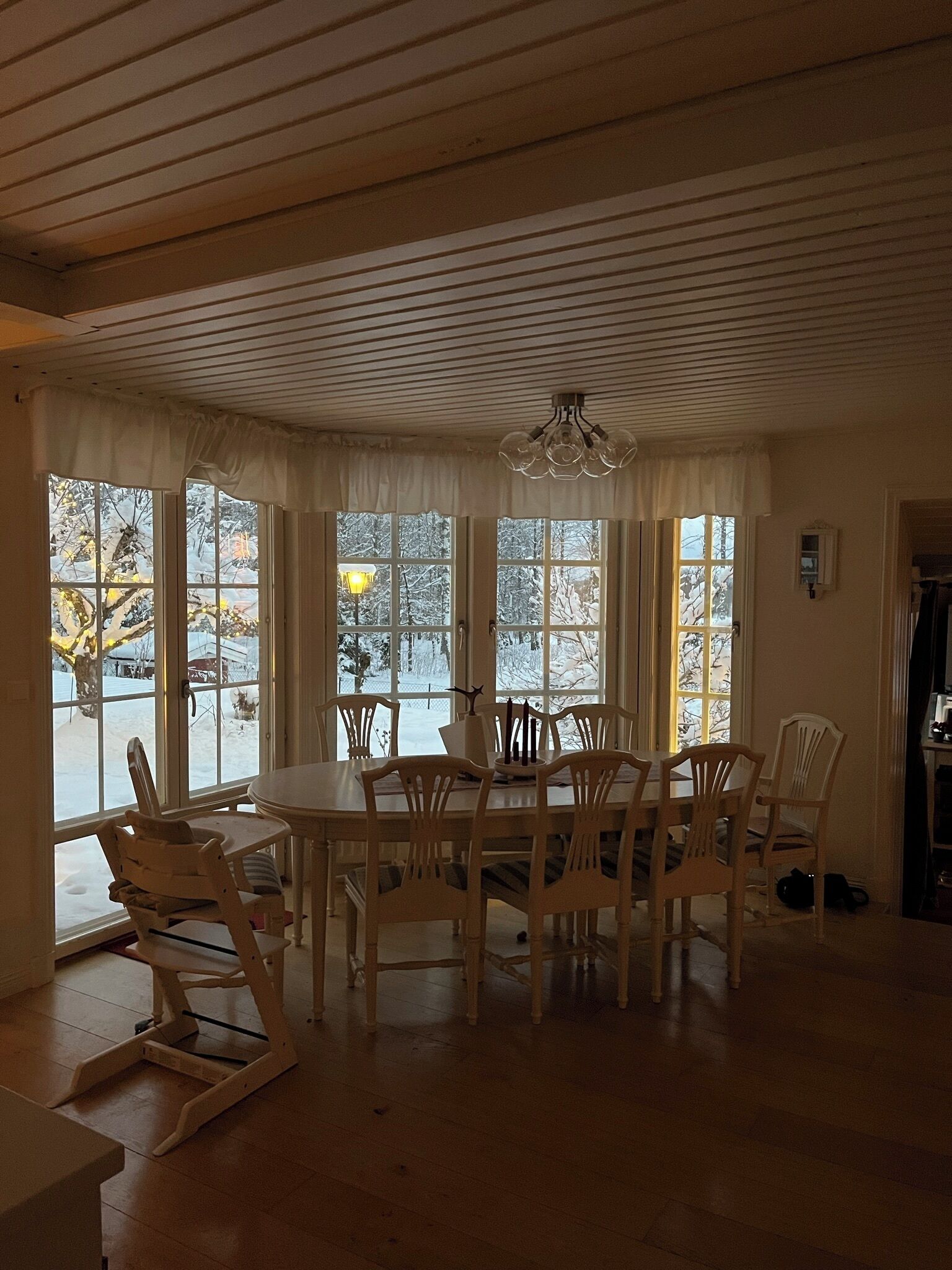 A dining area with a round table and six chairs, large windows with curtains, and a ceiling with wooden beams.