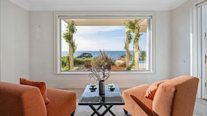 A room with a large window overlooking a garden and the sea, featuring two orange sofas and a black table with decorative items. - The Bay Crest Manor Views Pool Beach (Mount Eliza)