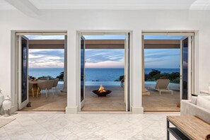 A modern living room with a large glass door leading to a patio with a fire pit and outdoor seating. - The Bay Crest Manor Views Pool Beach (Mount Eliza)