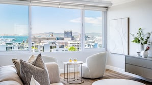 A modern living room with a large window offering a city view. There are two white armchairs, a small round table with two glasses of orange juice, and a white sofa with decorative pillows. - Charming Central Apartment Aquene bay (Cape Town)