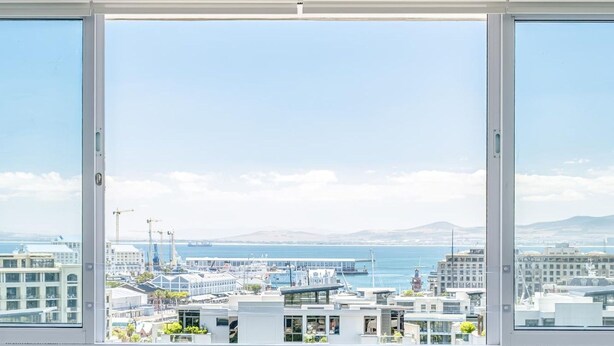 A modern living room with two sofas, a round coffee table with a fruit bowl, and a view of a cityscape through large windows. - Charming Central Apartment Aquene bay (Cape Town)
