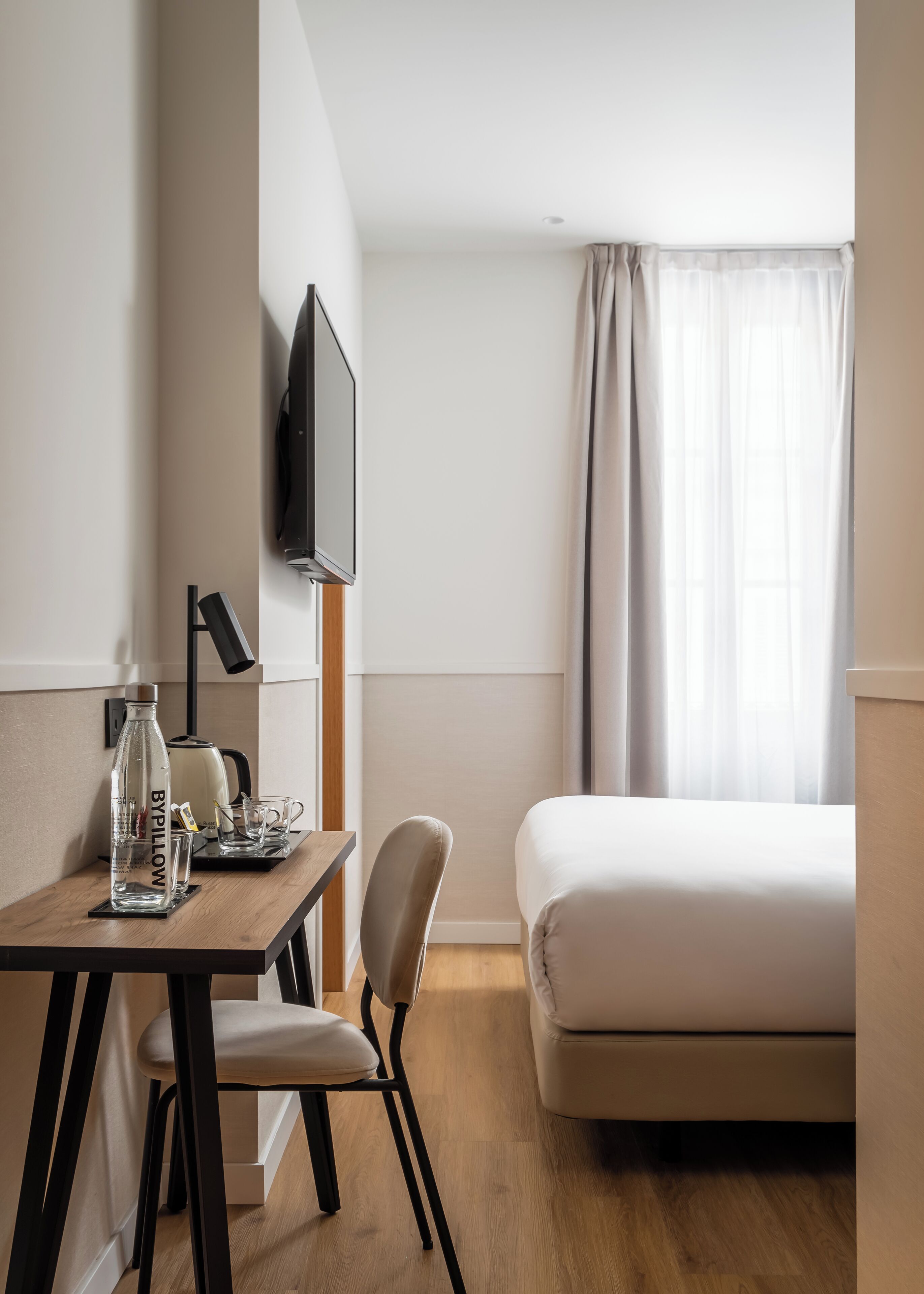 Una habitación de hotel moderna con un escritorio de madera, una silla, una cama, un televisor de pantalla plana montado en la pared y una ventana con cortinas.