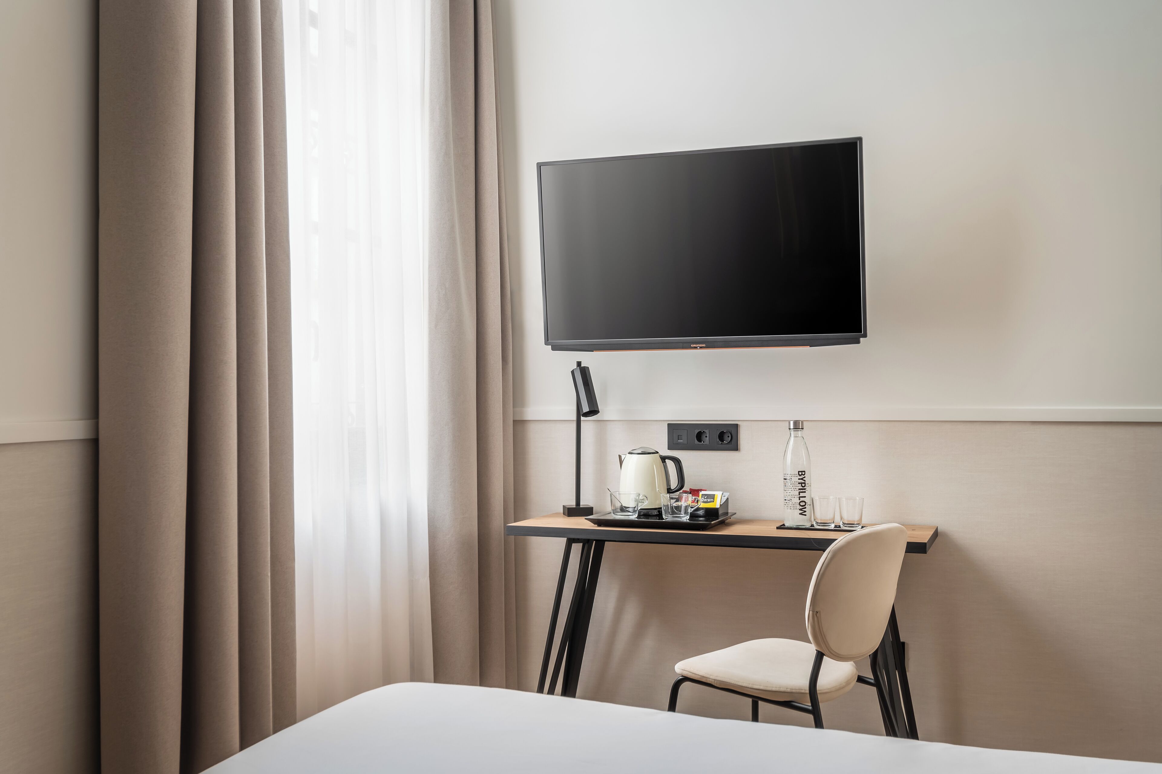 Habitación de hotel con cama, escritorio, televisor de pantalla plana, silla, tetera y botella de agua.