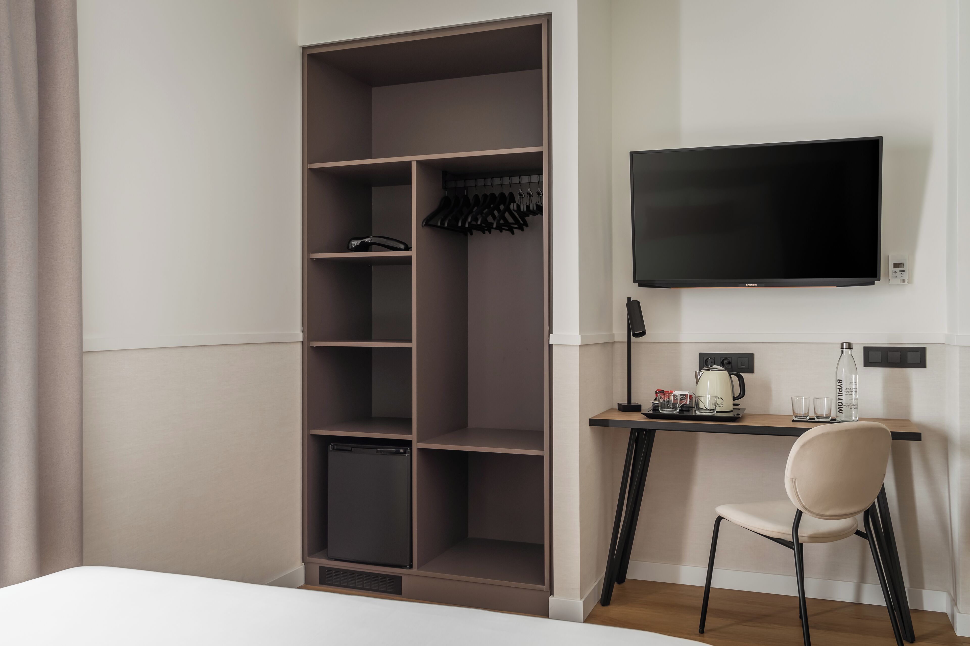 A hotel room with a wardrobe, a desk with a chair, a flat-screen TV, and a small refrigerator.