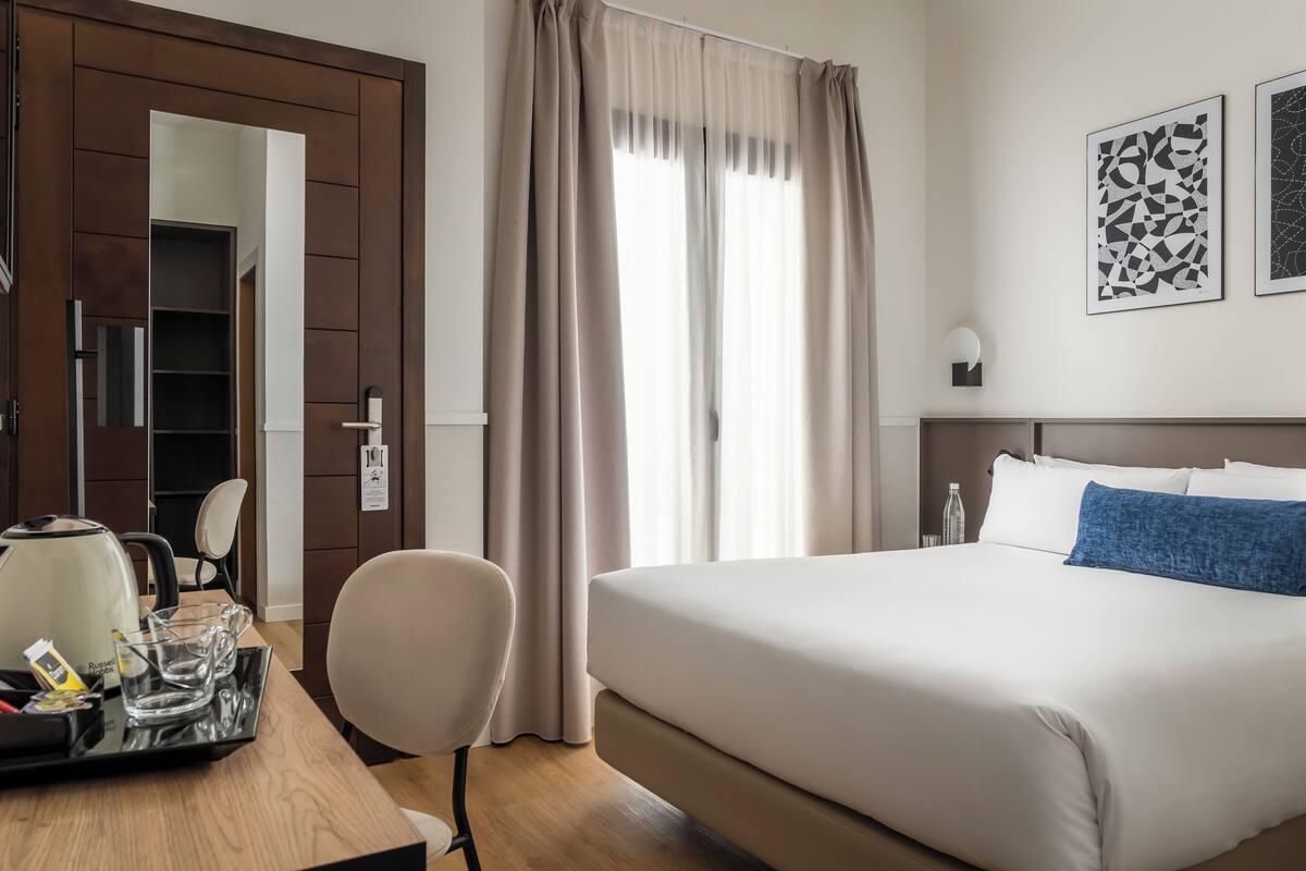 A hotel room with a bed, a desk with a kettle and glasses, a chair, and a wardrobe.