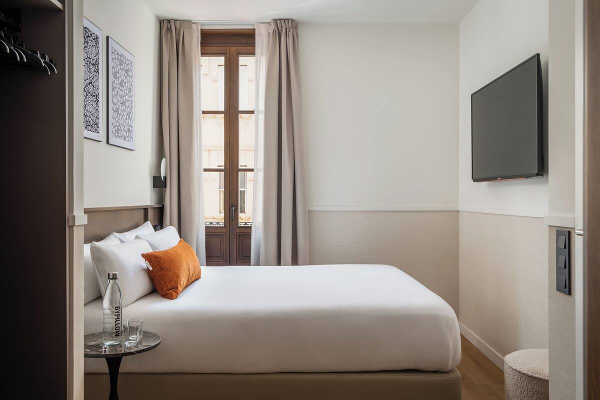 A hotel room with a bed, a small table with a bottle and glass, a TV, and a window with curtains.