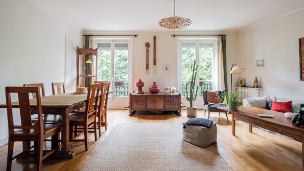 A spacious living room with a wooden dining table, chairs, a sofa, and a bench. There are two windows with curtains, a hanging light fixture, and a potted plant. - Boho on the Boulevard, by Veeve (Paris)