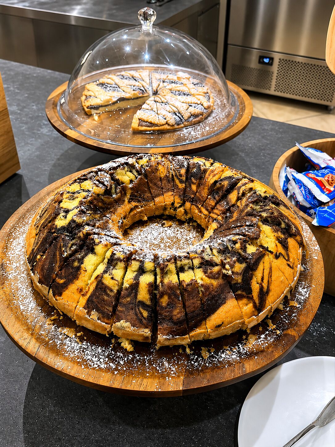 Un pastel bundt sobre una tabla de madera, un pastel cubierto en un soporte, y un microondas al fondo.
